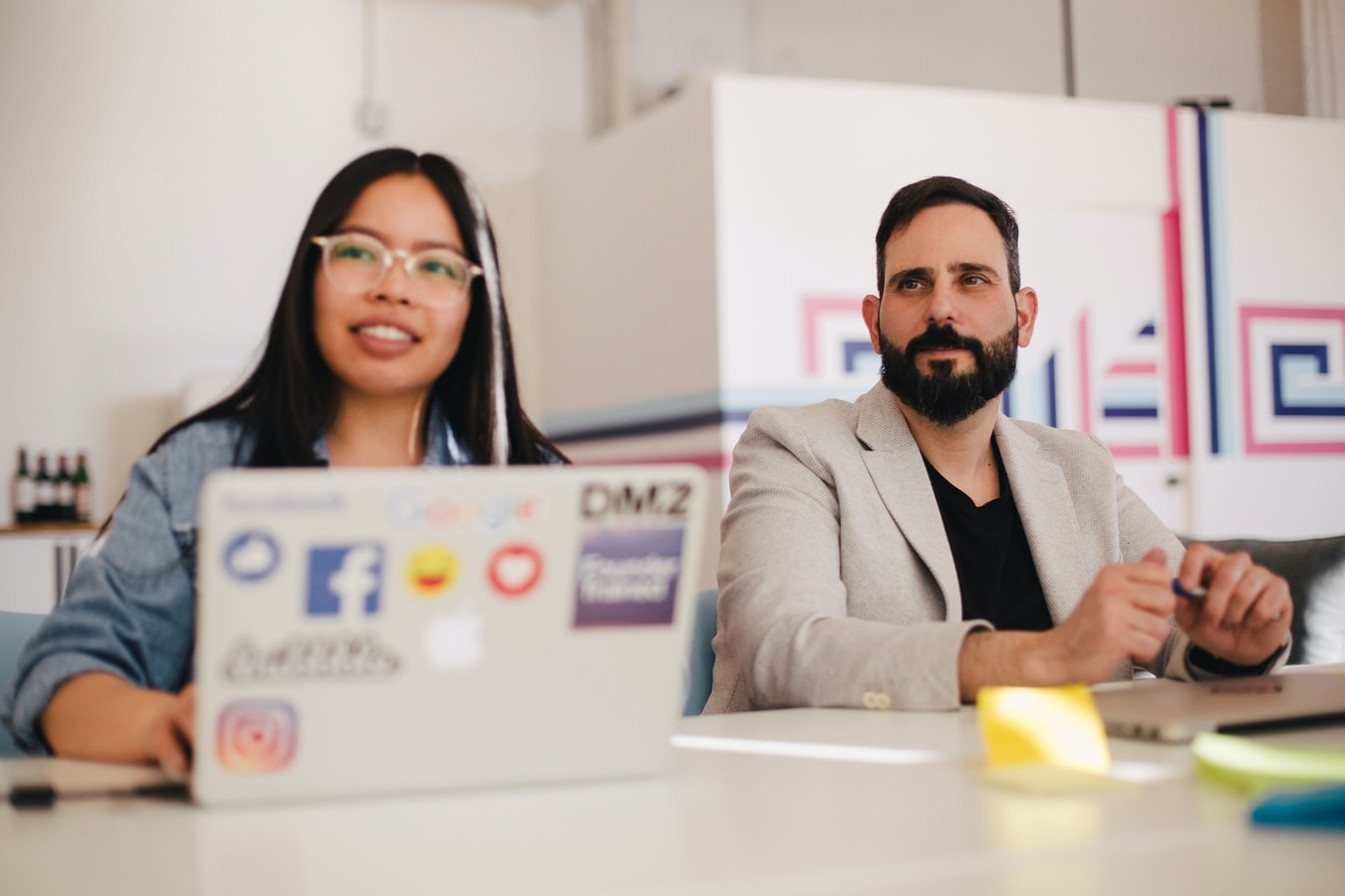 Two people sit at a table with laptops having a discussion